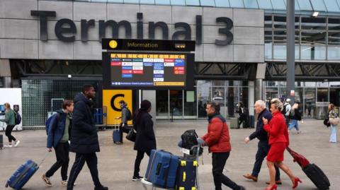 Passengers push luggage outside Terminal 3 as a gate information screen can be seen in the background