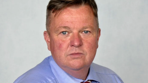 A man in a blue shirt and tie in front of a grey background. 