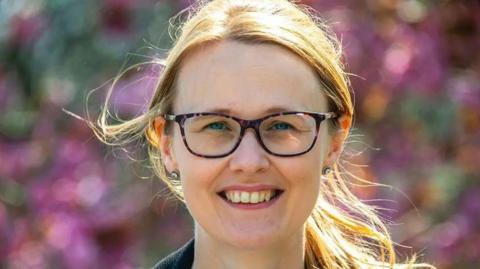 Cat Smith MP with long blond hair in a pony tail wearing glasses with purple rims with flowers blurred out in the background. She is smiling.