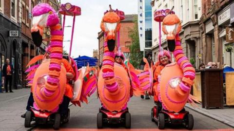 three flamingo puppets driven by people on mobility scooters on the streets at a previous festival