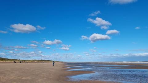 Bright blue sky with a few white clouds over a beach with the sea to the right 