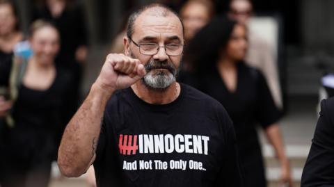 Malkinson, who has a beard and wears glasses, holds up his hand in a fist pump. He wears a black T-shirt with writing on it that reads: hashtag innocent and not the only one...