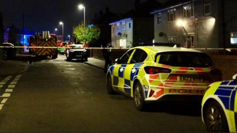 Two police cars parked on a street nearest the camera behind police tape across a road. In the distance a fire engine is parked up with blue lights on. There are houses either side and it's night time.