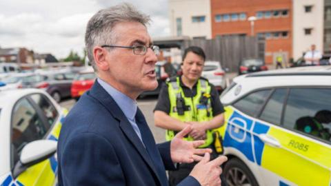 A man wearing a navy suit and tie is standing side-on, next to two police cars in a car park. A police officer is standing behind him. The man is talking and gesturing with his hands out in front of him. There are red brick buildings in the background. 