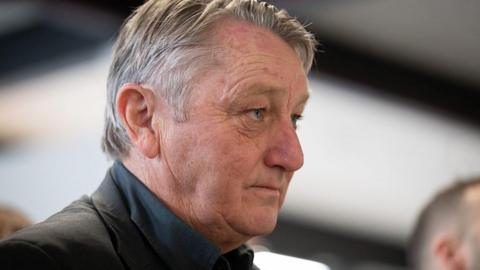 Anthony Slaughter with a dark suit and dark green shirt looking to the right of the picture. He has grey hair.