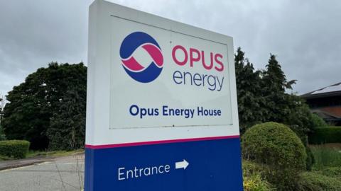 Large blue and white entrance sign for Opus Energy House, surrounded by trees and shrubs