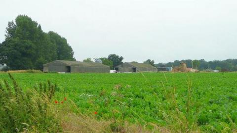 A view of farm buildings as seen from across a green field on a cloudy day.