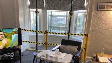 The living room, which has black and yellow tape cordoning off a bay window, there are scaffolding poles that seem to be propping up the ceiling, an arm chair is just in front with a small table, and a television is on in the corner.