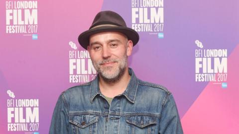 A man in a hat and open neck shirt, smiles at the camera at a film awards ceremony