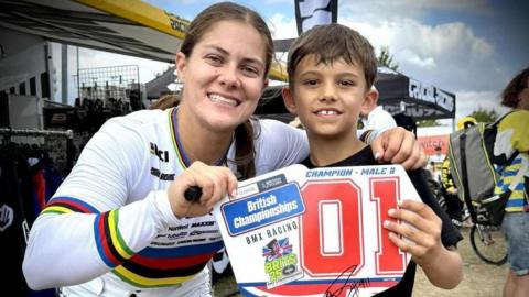 Archie Fisher is on the right of picture, posing with Team GB rider Beth Shriever. Beth is wearing a white racing suit. They are holding a sign saying 'British Championships BMX Racing Champion Male 8', a title Archie won at the British BMX Championships in Kent in 2025.