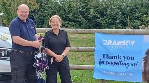 David Cobb, of Cobbs Country Store, dressed in a blue top and dark trousers standing in front of a field railing and blue Bransby Horses sign holding the purple collars. Next to him is PC Irving, who is smiling and wearing dark clothing. 
