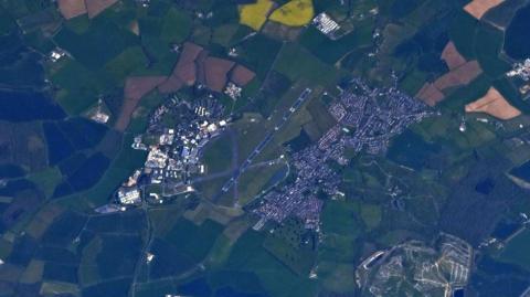 A photo taken of Cranfield from high above, you can see houses, farmer's fields and university buildings surrounding a runway.