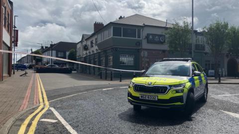 A marked police car is in the foreground. White police tape is across the street. In the background you can see buildings on either side of the street and double yellow lines along the left hand side as far as the eye can see.