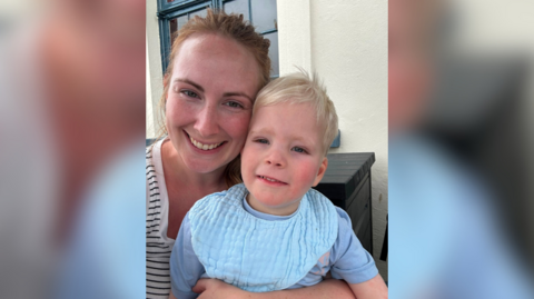 The image shows a woman holding her son while smiling. Her son is wearing a baby blue top and the woman is wearing a black and white striped top.