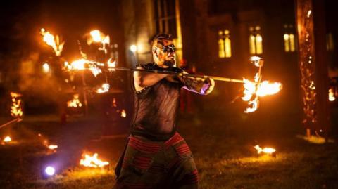 A man wearing warrior-style clothes and make-up holds a long stick, with fiery torches at either end. More torches can be seen on the grass outside a medieval church. The man has a Mohican-style haircut and short beard on his chin, wears black and white make-up around his eyes, and wears a black vest, resembling chainmail, and black and red trousers.