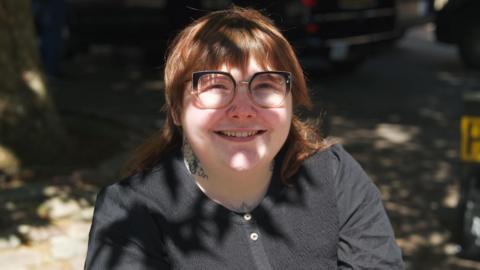 Rebecca Scollay sitting outdoors in her wheelchair. She is wearing a black long-sleeved T-shirt and is looking up at the camera. The sun is on her. The trees behind her are casting a shadow. She has auburn hair with a fringe and is wearing glasses.