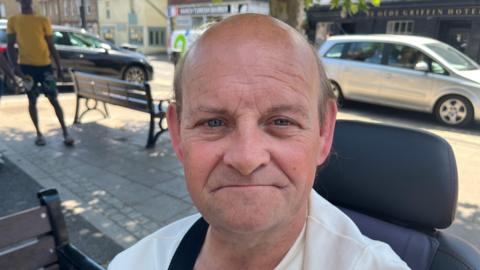 Christopher Smalley, 54, has a mainly bald head, with some dark hair on the sides of his head. He is sitting in a mobility scooter and behind him is a town square, with benches and cars passing on the nearby roadway.