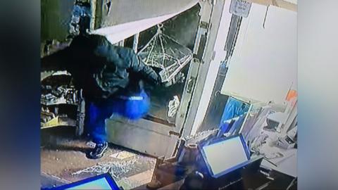 Man in black jacket, with hood up, and blue trousers breaks through the glass front door of a shop. He appears to be stumbling forward. Two blue screens are in the foreground and there is broken glass on the shop floor. 