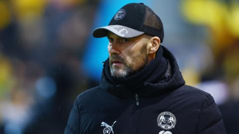 Peterborough United boss Luke Williams looks ahead wearing clubv winter coat and baseball cap
