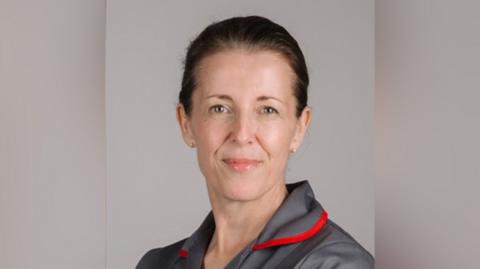 Sam Young  smiling in a grey and red nurse outfit. She has brown hair tied into a bun.