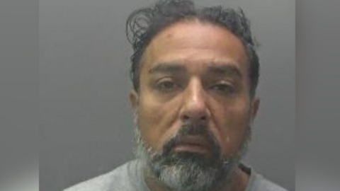 Chaudhry's custody image wearing a grey T-shirt. He has dark facial hair. 
