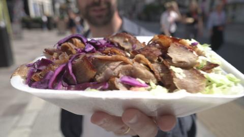 A man who is out of focus holding a kebab with brown meat, green lettuce and red (purple) cabbage. The packaging is white.