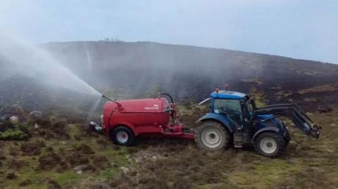A red slurry tank attached to the back of a tractor sits on a moorland spraying water across an area of burnt land