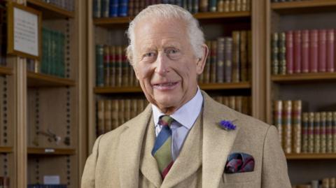 King Charles is wearing a beige suit. He stands in front of a collection of books in a library and is smiling at the camera