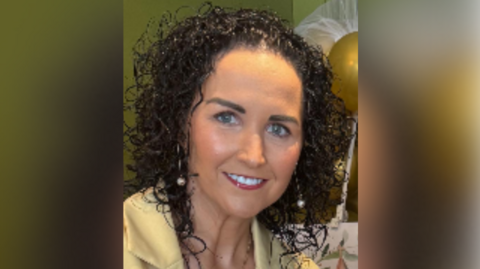 A woman with long black curly hair smiles at the camera as she wears a yellow blazer and gold necklace. She has blue eyes and is wearing make up. She is wearing gold dangly earrings with pearls on them.