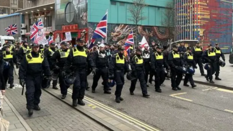 A small group of protesters with British and English flags march with a larger police escort