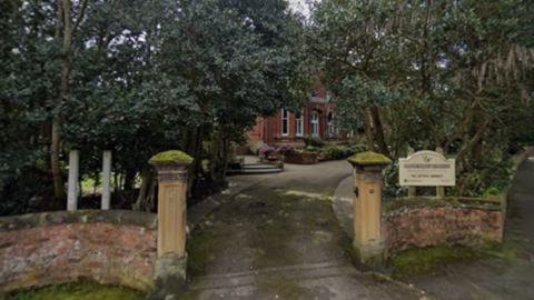 The front entrance of Dalemain House. It is a Victorian-era red-brick villa with trees surrounding its driveway. 