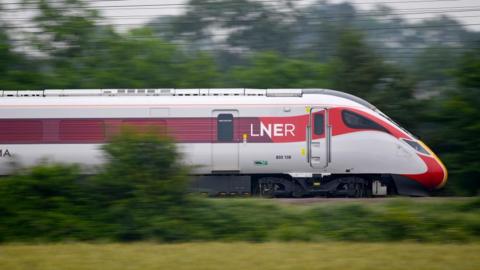 A high-speed train, with a sleek, streamlined front cab, passing through countryside. It is a red and white and says LNER on the side. Green grass and trees and electricity lines look blurred in the background, demonstrating the speed at which the train is travelling.