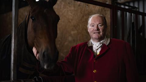 A huntsman in red jacket looks at the camera with his horse