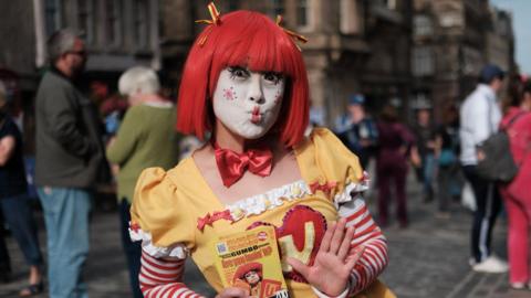 A doll-like clown in a yellow and red dress, bright red wig and doll clown make-up poses with a kiss while holding flyers advertising her show