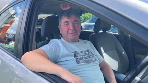 John Moreton is sitting inside his car. The window is down and he is looking at the camera, wearing a blue t-shirt. 