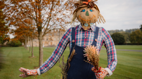 A scarecrow with a red and blue checked shirt and blue dungarees, with colourful gardening gloves and holding a bunch of dried flowers