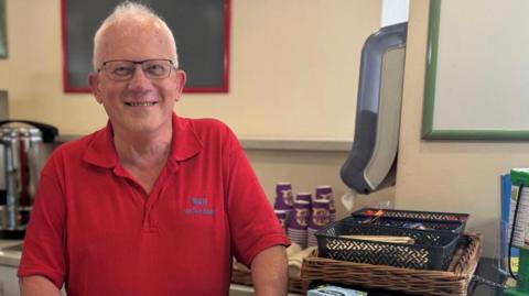 Gavin Stoppel with short white hair and glasses, wearing a red tee-shirt, standing in front of a stack of paper cups, a tea urn, tea bags and stirrers in a plastic tray