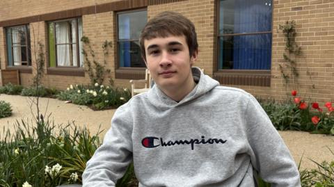 A teenage boy with short brown hair is pictured in a garden wearing a grey hoodie with the brand Champion written on it. 