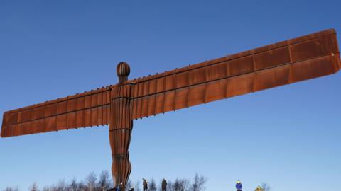 Antony Gormley's Angel of the North sculpture in Gateshead. The tall brown sculpture, which resembles a man with rectangular wings, stands tall over the wiry trees below.