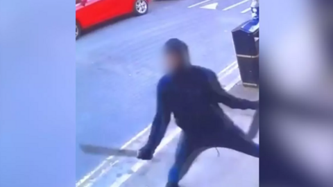 A CCTV image of a man with a blurred face, in dark clothing, wielding a machete in his right hand