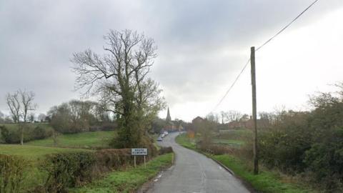 Streetview image Walcote Road, South Kilworth, showing a narrow, rural road leading to a village with a spire visible