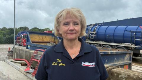 A woman with blonde hair smiles at the camera , she wears a polo shirt bearing the name of Pellows. Behind her is a facility where septic tank waste is processed. 