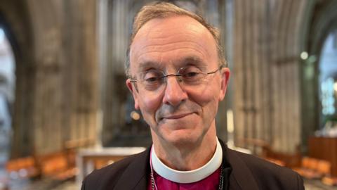 Bishop Inge wearing a purple shirt, dog collar and black jacket poses for camera.