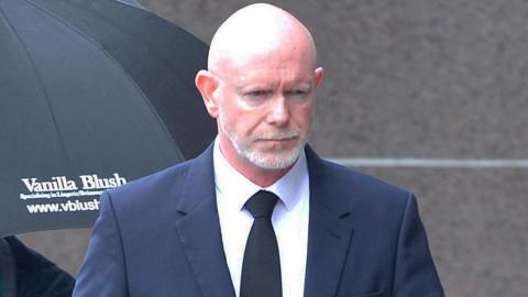 Philip Braat, a bald man with a well-trimmed grey beard and moustache, walking towards a court building. He has a serious expression and is looking at the ground. Braat is wearing a navy suit, white shirt and a black tie