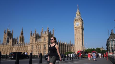 A woman walks along the pavement with the Houses of Parliament behind her. The sky is blue and the weather sunny.