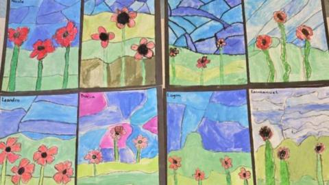 Artwork created by young children and used to decorate a wall shows different pictures of poppies in fields under a blue sky.