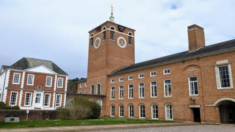 County Hall in Exeter, the base of Devon County Council. It consists of a large brown brick built building with a clock on the tower of one of the buildings. There is a weather vane on top of the tower.