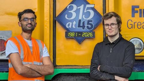 Lakshman Pathak and Brad Wright stand in front of the end of a Class 315 electric train. The front of the train is yellow with a blue square that reads 315 at 45, 1981-2026. Lakshman has dark hair, glasses and wears a white T-shirt with an orange hi-vis bib over the top. Brad has dark blonde hair, glasses and wears a black jumper. Both men have their arms crossed in front of their torsos and are slightly smiling. 