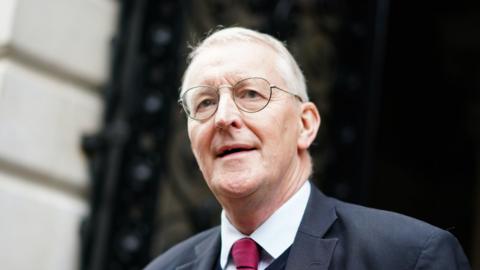 Hilary Benn in a grey suit jacket and red tie, white shirt, with glasses, smiling against building