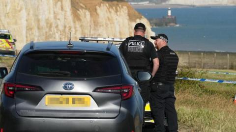 Two police officers stood next to a car in a field. The field overlooks a cliff edge, with sea in the background and a lighthouse on a jetty.